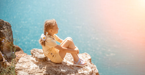 Happy girl perched atop a high rock above the sea, wearing a yellow jumpsuit and braided hair,...