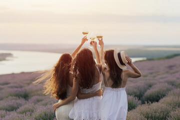 Lovely ladies drinking wine in lavender violet field at sunset. Summer happy mood. Girlfriends relaxing on summer sunset with river on the background.	
