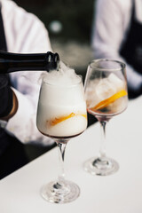 Bartender pours a sparkling wine in glass. Two glasses of cocktails on the table in the bar. 
