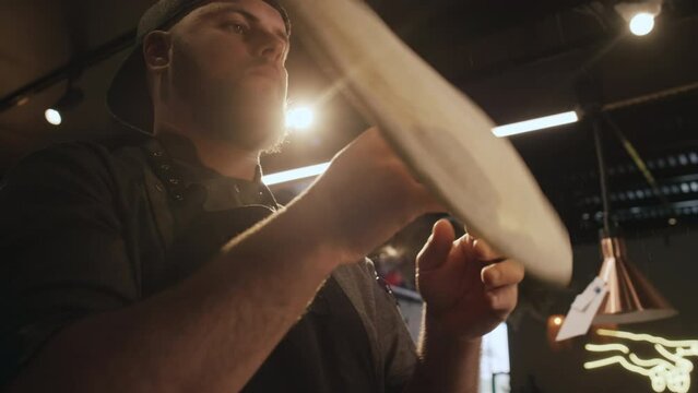 Medium low angle slowmo shot of bearded professional Caucasian male chef working in dimly lit restaurant kitchen and throwing, stretching and spinning dough into thin round shape