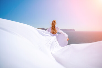 Fototapeta na wymiar A blonde woman with long hair stands on a sunny seashore in a flowing white dress, with the silk fabric waving in the wind, and blue sky and mountains in the background.