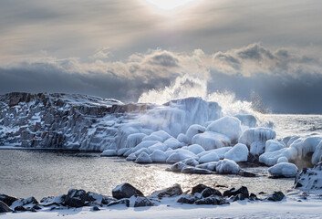 Waves crash over ice covered rocks while sea smoke hovers over the water on a cold winter day