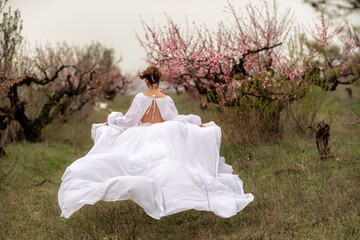 Fototapeta na wymiar Woman peach blossom. Happy curly woman in white dress walking in the garden of blossoming peach trees in spring