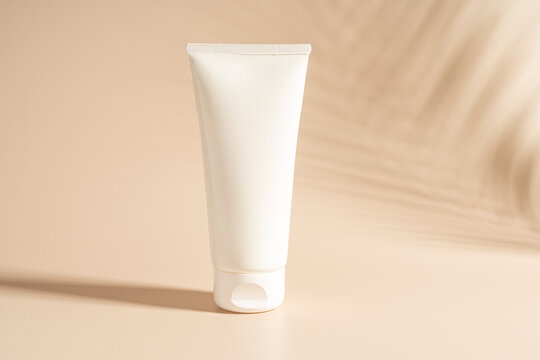 A white tube of face cream standing on a beige background with shadows nearby