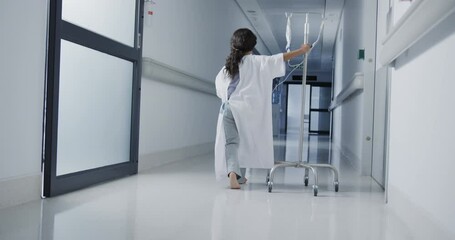 Biracial sick girl patient walking with drip in hospital corridor in slow motion