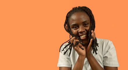 Portrait of young African woman looking at camera with big smile, playing with her hair with hands against orange background