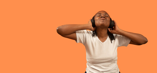 Portrait of African young woman listening to music with headphones, eyes closed, head up and holding headphones with hands against orange background