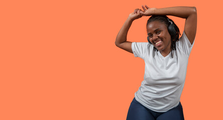 Portrait of happy young African woman dancing and listening to music with headphones, arms up, dressed in white t-shirt and blue pants against orange colored background