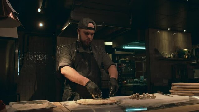 Medium shot of Caucasian man in chef uniform, black apron and cap making pizza alone in dimly lit restaurant kitchen, sprinkling handfuls of grated cheese on dough, then flattening and spreading it