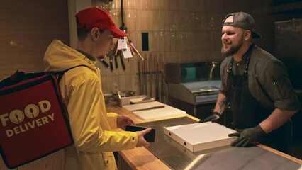 Medium shot of delivery service courier with bag walking into restaurant, showing order number on smartphone, smiling chef packing fresh pizza into box, giving to him, and courier thanking and leaving