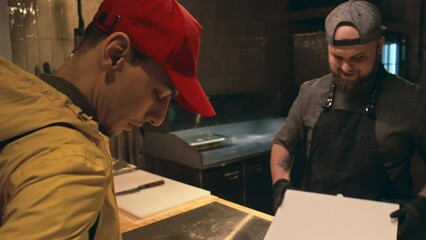 Medium follow shot of food delivery service courier with backpack walking into restaurant, showing order to male chef, who is packing pizza in cardboard box and giving away, both smiling and chatting - Powered by Adobe