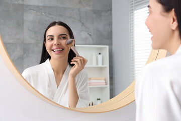 Beautiful young woman applying makeup with brush near mirror in bathroom