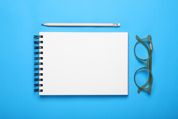 Open notebook, pencil and glasses on light blue background, flat lay
