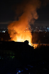 Fototapeta na wymiar Building on fire at night in city. Orange flames and heavy smoke pouring out of burning damaged house during nighttime. Fire hazard in buildings concept