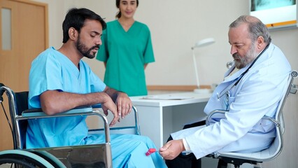 Asian senior male doctor treating knee pain to an Indian male patient sitting in a wheelchair in the treatment room at the hospital