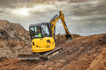 Mini excavator at the construction site on the edge of a pit against a cloudy blue sky. Compact construction equipment for earthworks. An indispensable assistant for earthworks. © Anoo