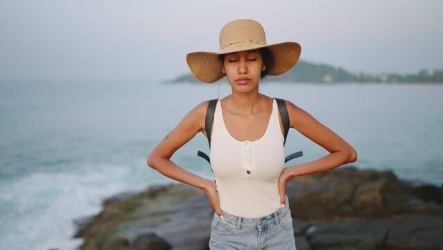 Pretty african woman opens eyes on camera, enjoys ocean view standing on island on sunrise. Biracial female in straw hat has emotional escape on travel to sea destination. Tourist bipoc girl relax.
