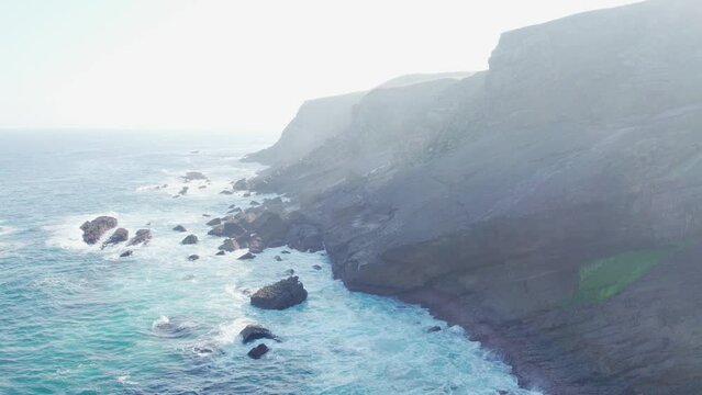 sea waves on ridges and peaks of mountain coastline of Playa de Tagle, Spain, cinematic aerial shot with sun flaring sunflare in a hazy sky.
