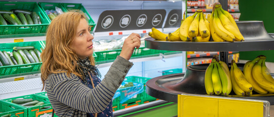 Mature pretty blonde woman worried and amazed at the high prices when buying bananas from a shelf...