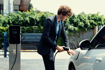 Progressive businessman with electric car recharging at public charging station at modern city...