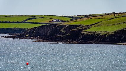 A house on a rocky Irish shore on a sunny summer day. Atlantic coast of Ireland, landscape. Green hills. Green grass field near body of water