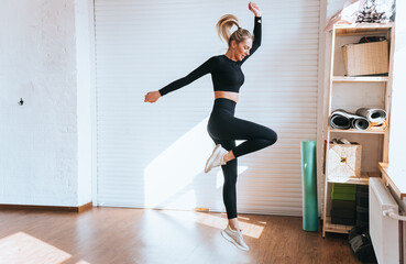 Fit caucasian young blonde woman in black sportswear jumps, training at fitness club on sunny day. Active Australian girl at exercise warms up before training with weights. Gym, sport club.