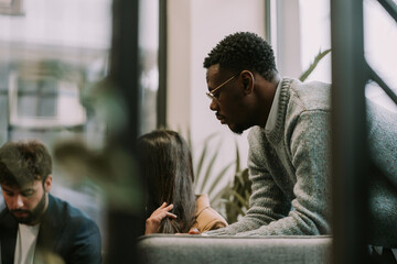 Black male person standing and leaning on sofa at the office. He is looking at his white coworkers working on the lap top