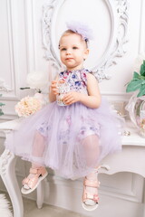 Baby girl elegant dress. A one-year-old girl in a puffy dress and a cute bow poses against the backdrop of a bright room with a dressing table and flowers.