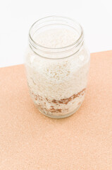 immagine con vista dall'alto di contenitore in vetro aperto con chicchi di riso e sfondo sfuocato in sughero su superficie bianca