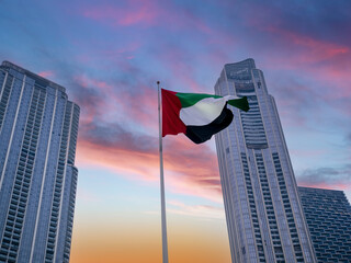 The flag of the United Arab Emirates is fluttering in the wind against the backdrop of skyscrapers at sunset as a concept of the symbols of Dubai