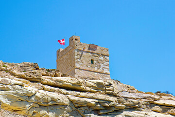Scenic view of rock with medieval stone tower and flag of Malta at Malta island on a sunny hot...