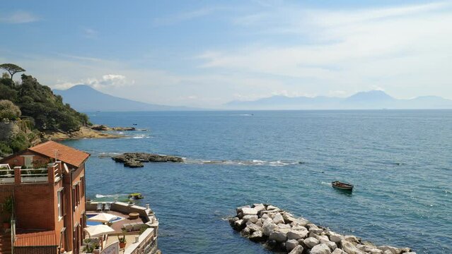 Naples, Italy. In Posillipo, on a sunny day, view of Marechiaro stretch of coast with a boat maneuvering into the bay. In the background Mount Vesuvius. Real time video. 2023-04-23.