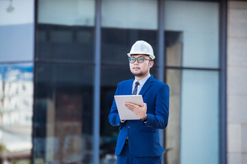 Handsome engineer wearing suit and hard hat or white helmet on building background. Engineer using tablet for checking construction about modern building. copy space for text.