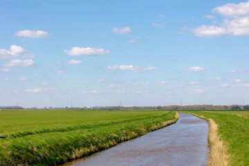 Fototapeta na wymiar Spring countryside landscape with flat and low land, Typical Dutch polder with green meadow under blue sky and white cloud, Small canal or ditch between the grass field, Drenthe province, Netherlands.