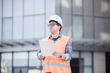 Handsome engineer wearing safety vest and white hard hat on building background. Engineer using Tablet for checking construction about modern building. copy space for text.