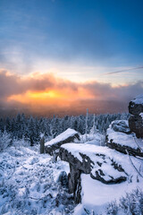 Dramatischer Himmel bei Sonnenuntergang mit bunten Wolken im Winter bei Schnee mit Felsen im Gebirge.