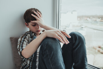 Close up lonely little boy, sitting at window alone, upset unhappy child waiting for parents, thinking about problems, bad relationship in family, psychological trauma