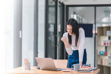 Obraz premium Happy and excited, a young asian woman student or office worker, celebrates achieving her goal of winning an online competition or receiving good news via email by raising hands in euphoria.