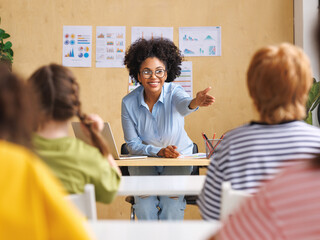  African american teacher smiling and looking at students and asks them  in class during lesson.