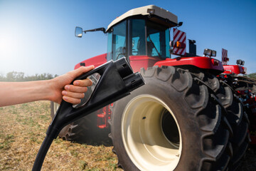 Hand with electric vehicles charging plug on a background of agricultural tractor. Concept	