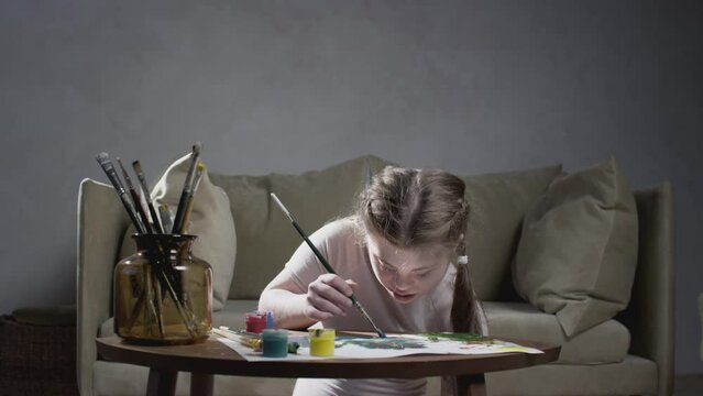 Portrait of a girl with Down Syndrome. The little girl draws with paints and a brush at home. Creates art and creativity. A person with special needs. Chromosomal genetic disorder in a child.