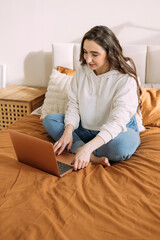 young curly girl in casual clothes works online from home on the bed in the bedroom