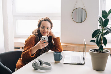 Smiling young woman texting on cellphone while working remotely at home office technology and...
