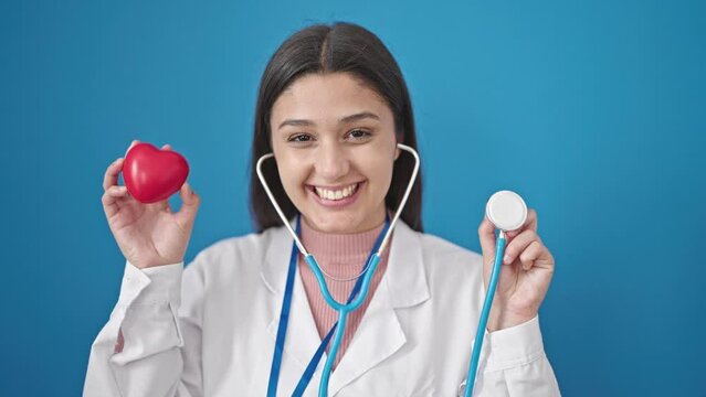 Young beautiful hispanic woman doctor smiling confident holding stethoscope and heart over isolated blue background