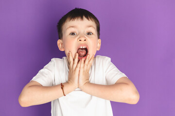Oh my god, wow! Portrait of funny amazed preschool boy looking at camera with shocked astonished expression and keeping hands on face, screaming in surprise studio shot isolated on purple background.