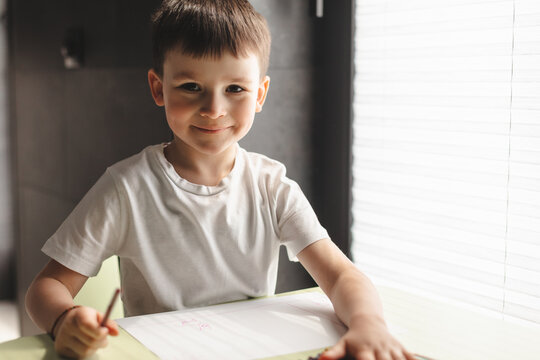 Little preschool boy in white t-shirt draw paint using color pencil and paper sitting at the desk in room at home. Children paint. Kids draw. Creative boy. Happy boy look at camera.