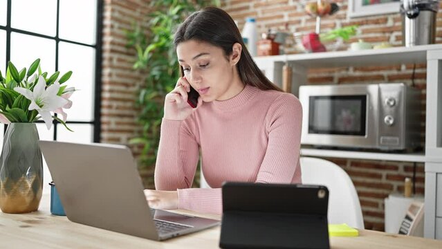 Young beautiful hispanic woman using laptop and touchpad talking on smartphone at dinning room