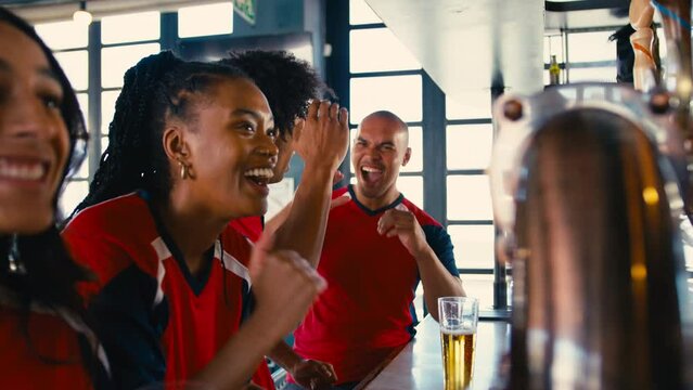 Multi-cultural group of friends wearing team shirts in sports bar celebrating as they watch game on TV together - shot in slow motion
