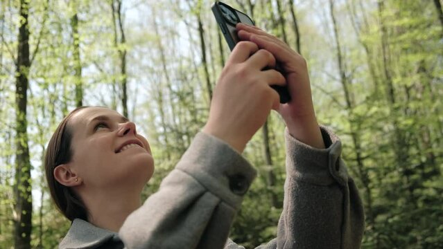 Happy young brunette woman using smartphone in spring forest. Traveler female hiker using phone. Cute girl hiker takes smart phone pic of beautiful woods. Concept adventure travel blogger wanderlust