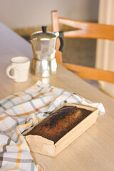 A chocolate cake, cupcake in an eco-wrap on a kitchen table. Rustic vertical still life with a checkered towel, a cup of coffee and a geyser coffee maker. Homemade pastries desserts in morning light.